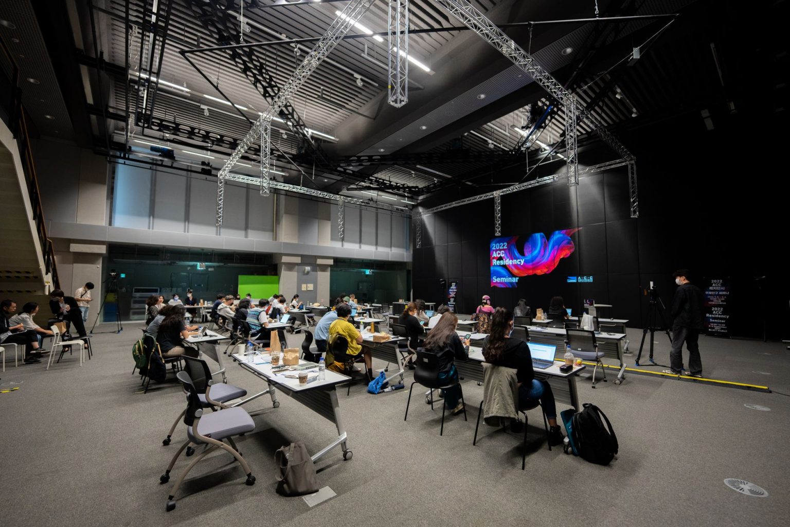 Participantes assistem a um seminário educacional em um espaço interno com telas e assentos.