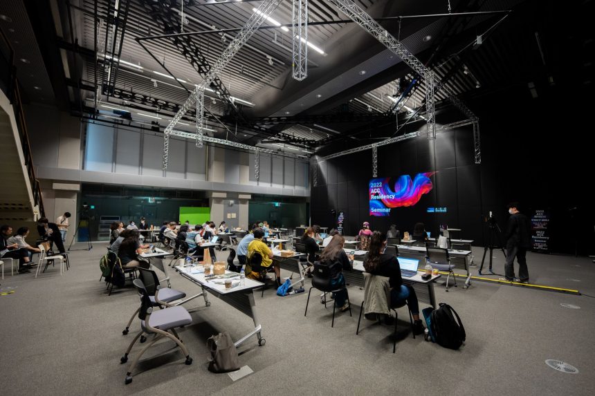 Participantes assistem a um seminário educacional em um espaço interno com telas e assentos.