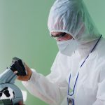 Cientista usando roupa de proteção examina amostras ao microscópio em um laboratório.