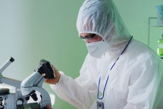 Cientista usando roupa de proteção examina amostras ao microscópio em um laboratório.