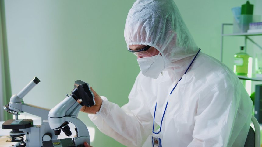 Cientista usando roupa de proteção examina amostras ao microscópio em um laboratório.