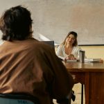 Professor conversa com estudante em uma sala de aula universitária.