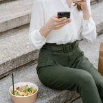 Mulher sentada em degraus, usando o celular ao lado de uma salada e de uma embalagem de papel.