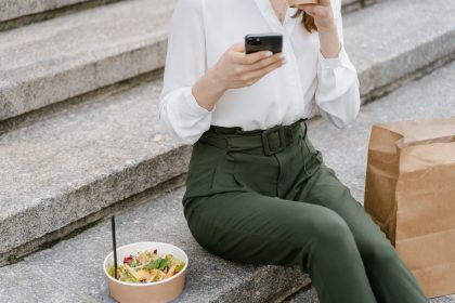 Mulher sentada em degraus, usando o celular ao lado de uma salada e de uma embalagem de papel.