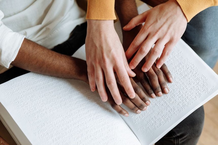 Duas pessoas leem juntas um livro em Braille sobre uma mesa.