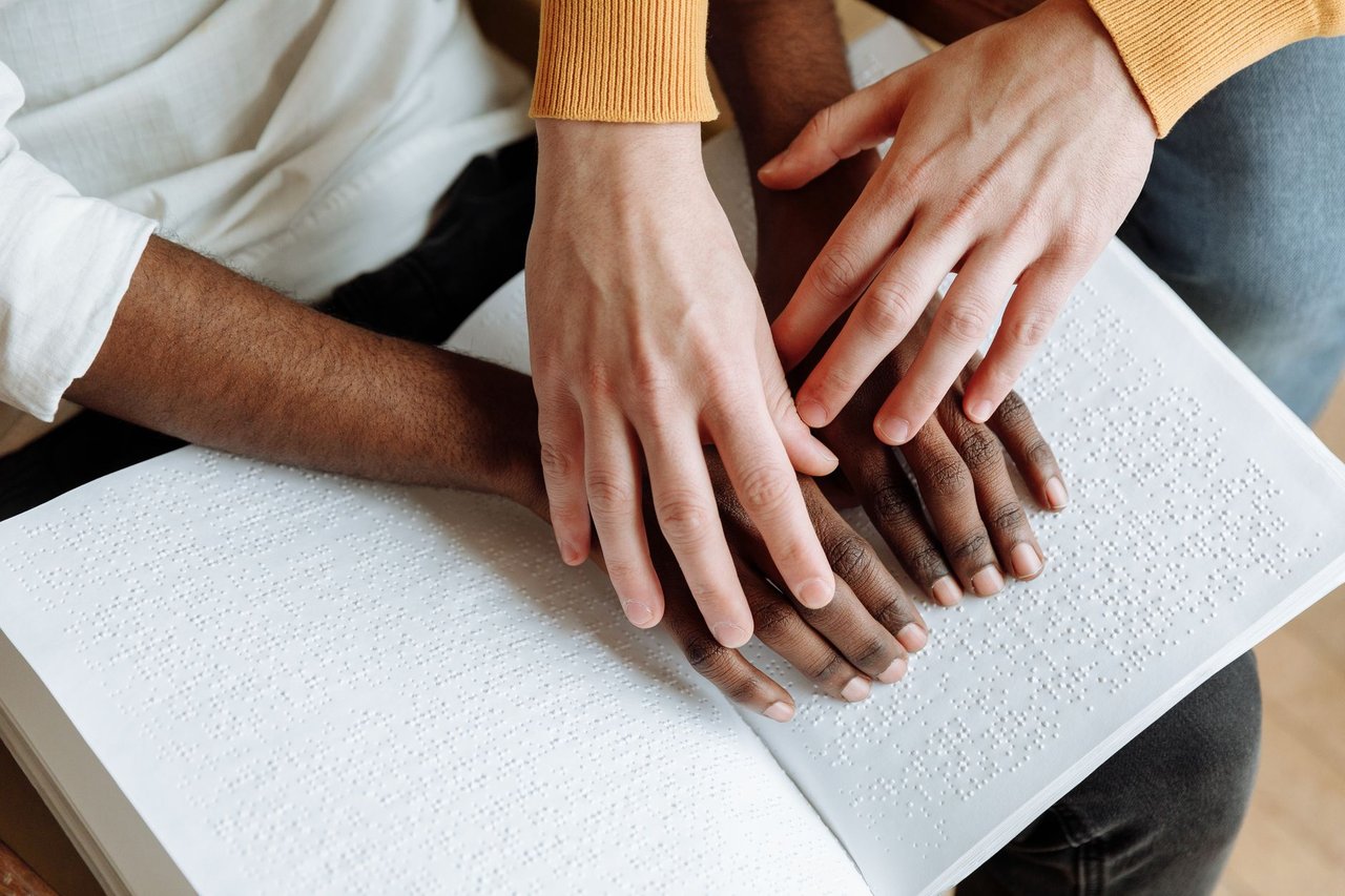 Duas pessoas leem juntas um livro em Braille sobre uma mesa.