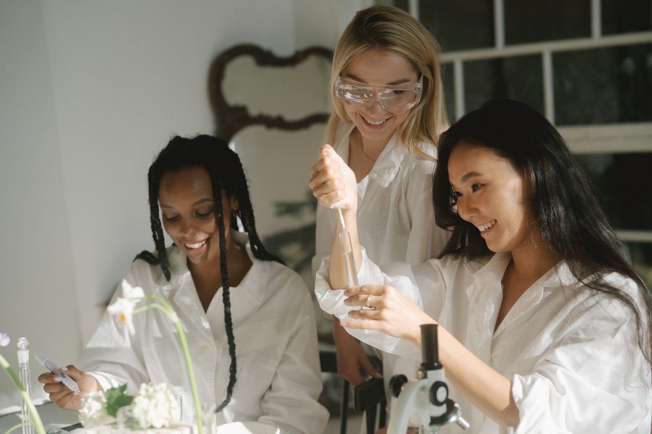 Três mulheres realizam um experimento científico com tubos de ensaio e equipamentos de laboratório.