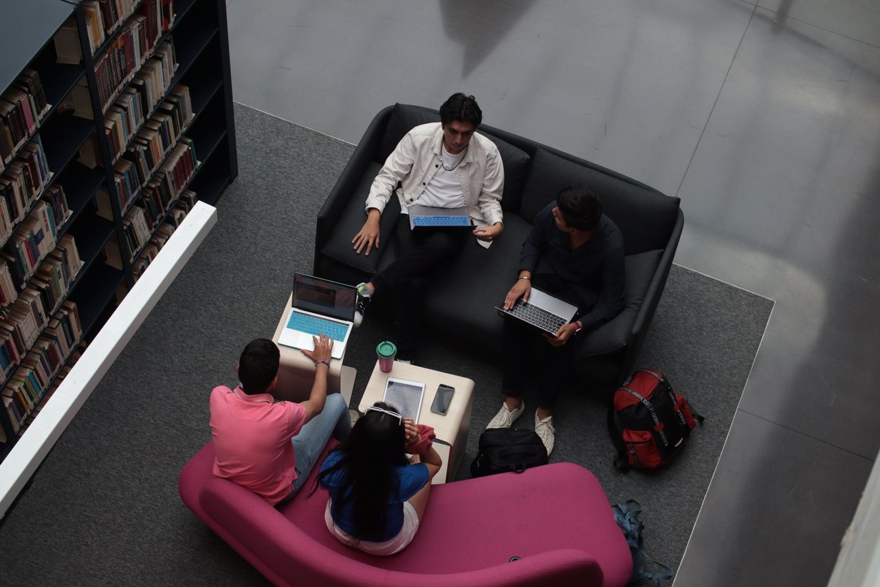 Grupo diverso de adultos colaborando em laptops na área de convivência de uma biblioteca