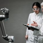 Pesquisadores de jaleco e óculos de proteção analisam um braço robótico em um laboratório.
