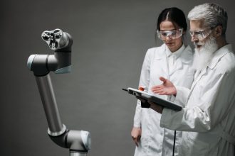 Pesquisadores de jaleco e óculos de proteção analisam um braço robótico em um laboratório.