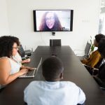Funcionários reunidos em uma sala de conferências participam de uma videochamada com um colega exibido em uma televisão.