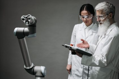 Pesquisadores de jaleco e óculos de proteção analisam um braço robótico em um laboratório.