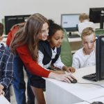 Professora orienta crianças durante uma aula de tecnologia em uma sala de aula moderna, diante de computadores.