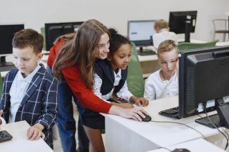 Professora orienta crianças durante uma aula de tecnologia em uma sala de aula moderna, diante de computadores.
