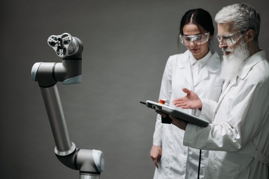 Pesquisadores de jaleco e óculos de proteção analisam um braço robótico em um laboratório.